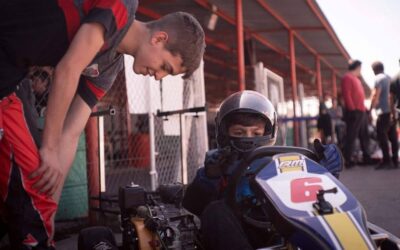 Jeune pilote en combinaison bleue et casque, assis dans un kart jaune et bleu, encadré par un adulte dans une zone de paddock
