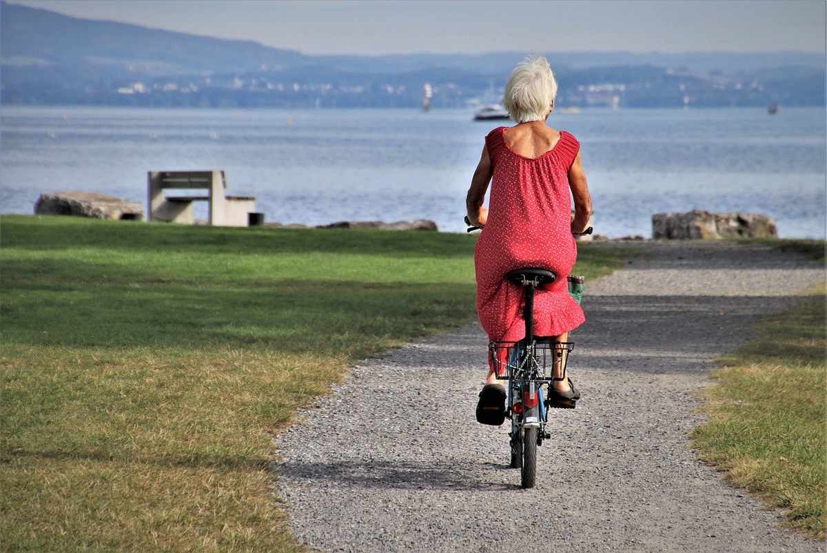 Femme en robe rouge pédale un vélo sur un chemin de gravier près d'un lac avec des montagnes en arrière-plan Femme en robe rouge pédale un vélo sur un chemin de gravier près d'un lac avec des montagnes en arrière-plan