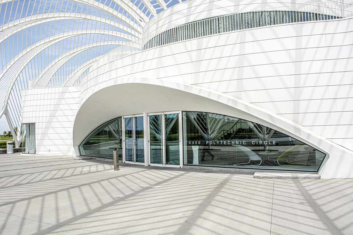 Bâtiment blanc aux lignes courbes, entrée vitrée, allée et ombres au sol